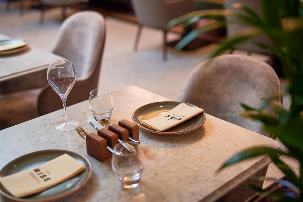 The Grand, York Hotel Exterior photo The photo shows a elegantly set dining table in a restaurant. The table has a light-colored marble surface and features two plates, each with a napkin that has the word "RISE" printed on it. There are also wine glasses and a water glass, along with u