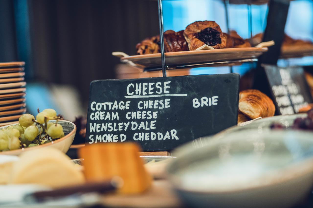 The Grand, York Hotel Exterior photo The photo displays a selection of cheeses and pastries on a catering table. A chalkboard sign lists various types of cheese, including cottage cheese, cream cheese, Wensleydale, smoked cheddar, and brie. In the background, there are baked goods, such
